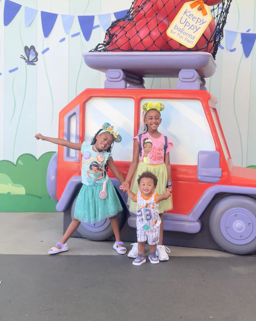 The sisters and their little brother smiling in front of a big red toy car on a family trip