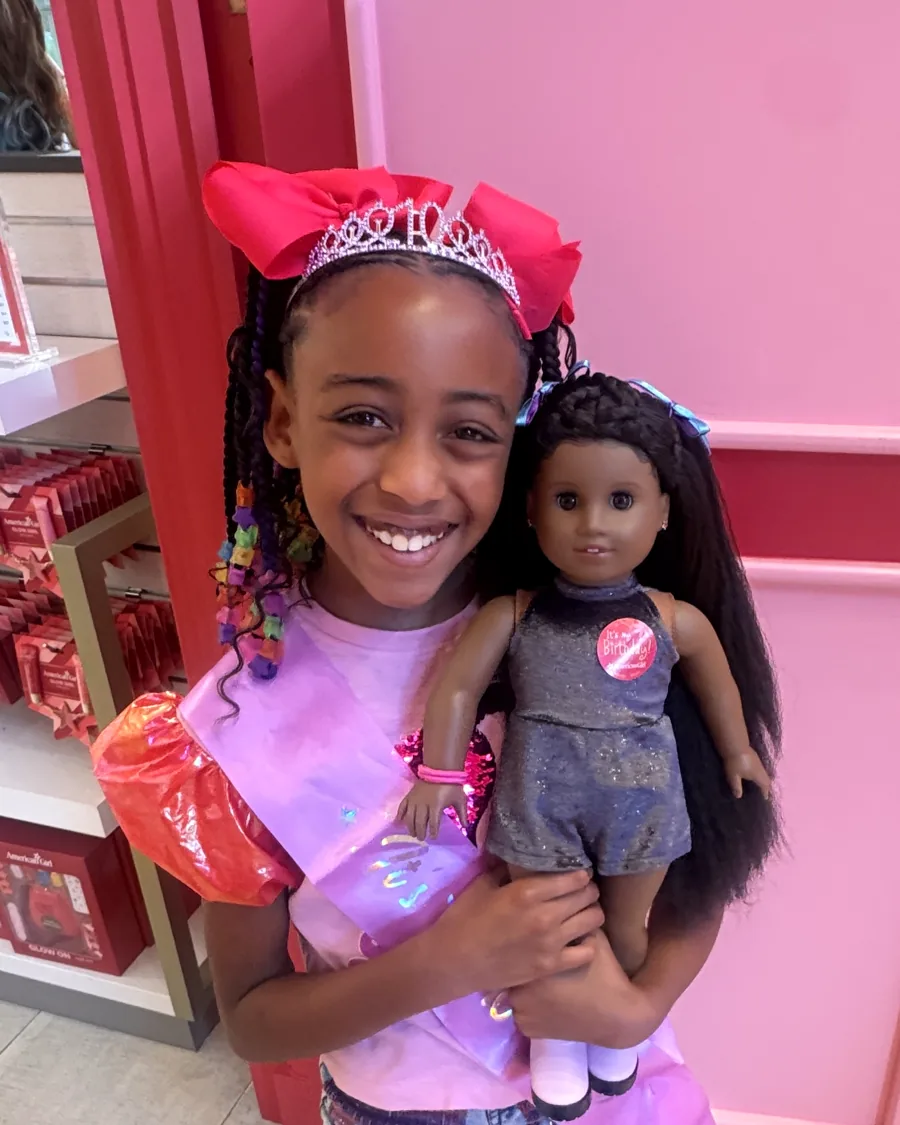 One sister smiling with her doll on a birthday outing, wearing a tiara and big pink bow